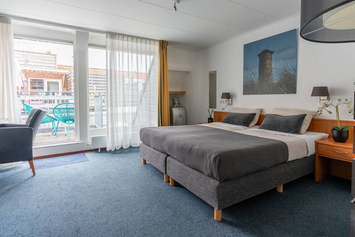 Ett hotellrum med en stor säng, ett skrivbord, en stol, ett fönster med gardiner och en tavla på väggen.