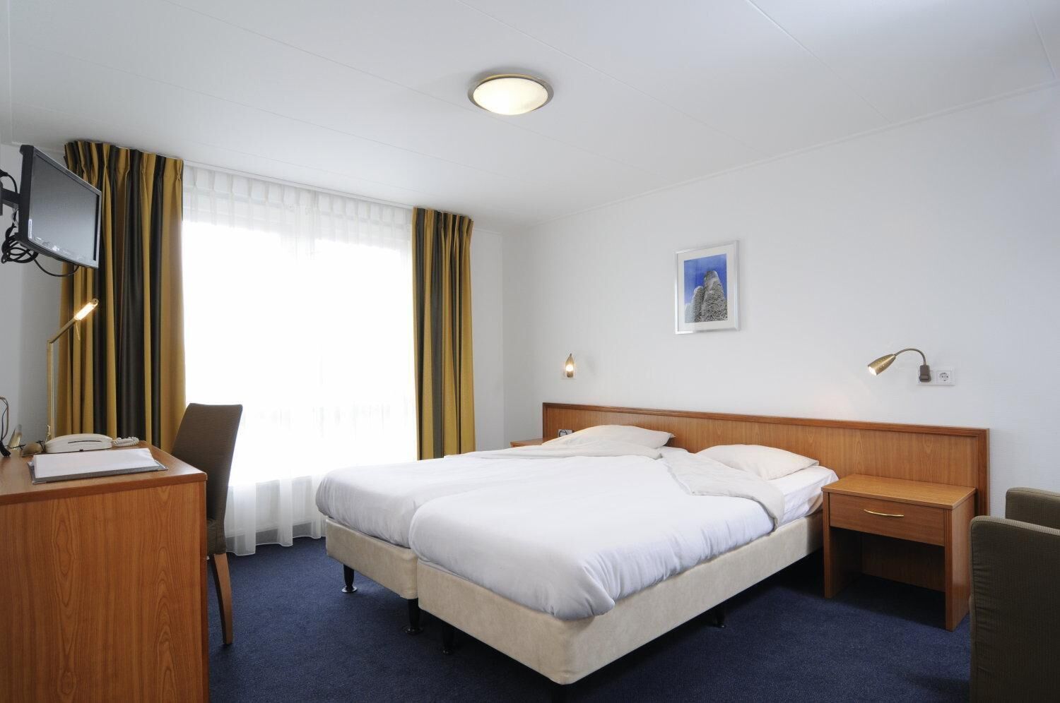 Une chambre d’hôtel avec un lit double, un bureau avec un téléphone, une chaise, une table de chevet avec une lampe et une fenêtre avec des rideaux.