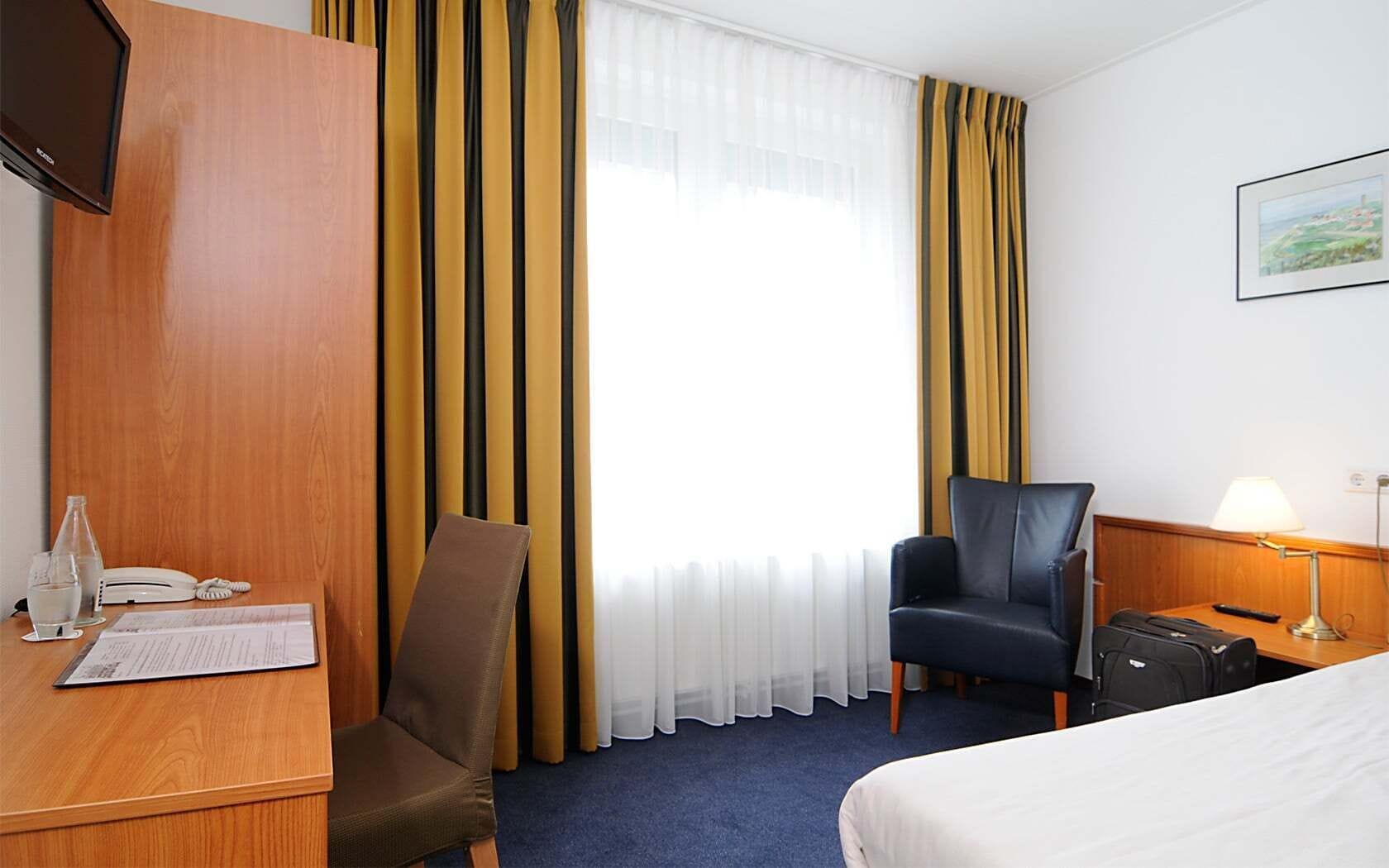 Ein Hotelzimmer mit einem Holztisch, einem Stuhl, einem Bett, einer Schreibtischlampe und einem Fenster mit Vorhängen.