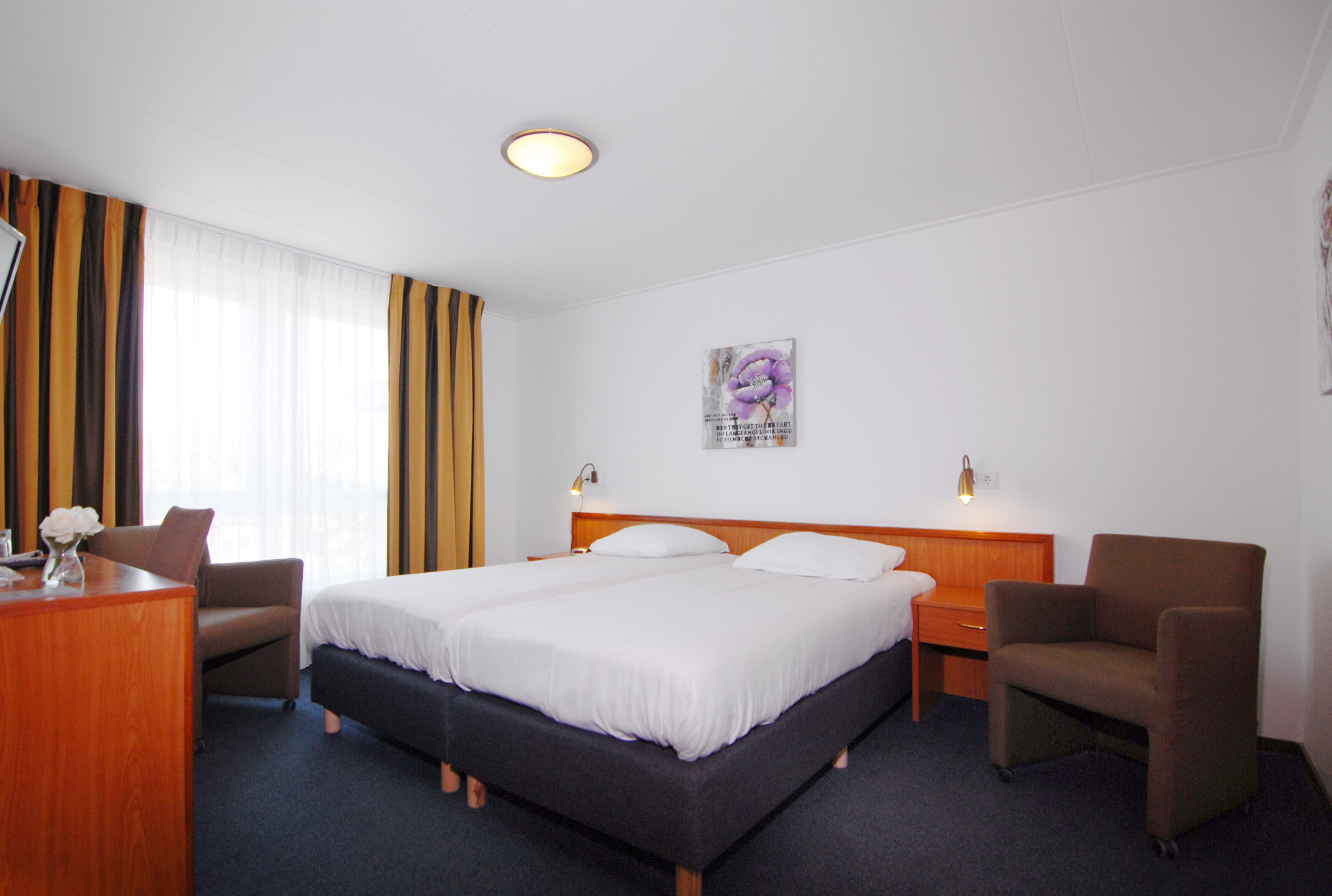 Quarto de hotel com uma cama grande, duas mesas de cabeceira, uma escrivaninha com cadeira e uma poltrona pequena.