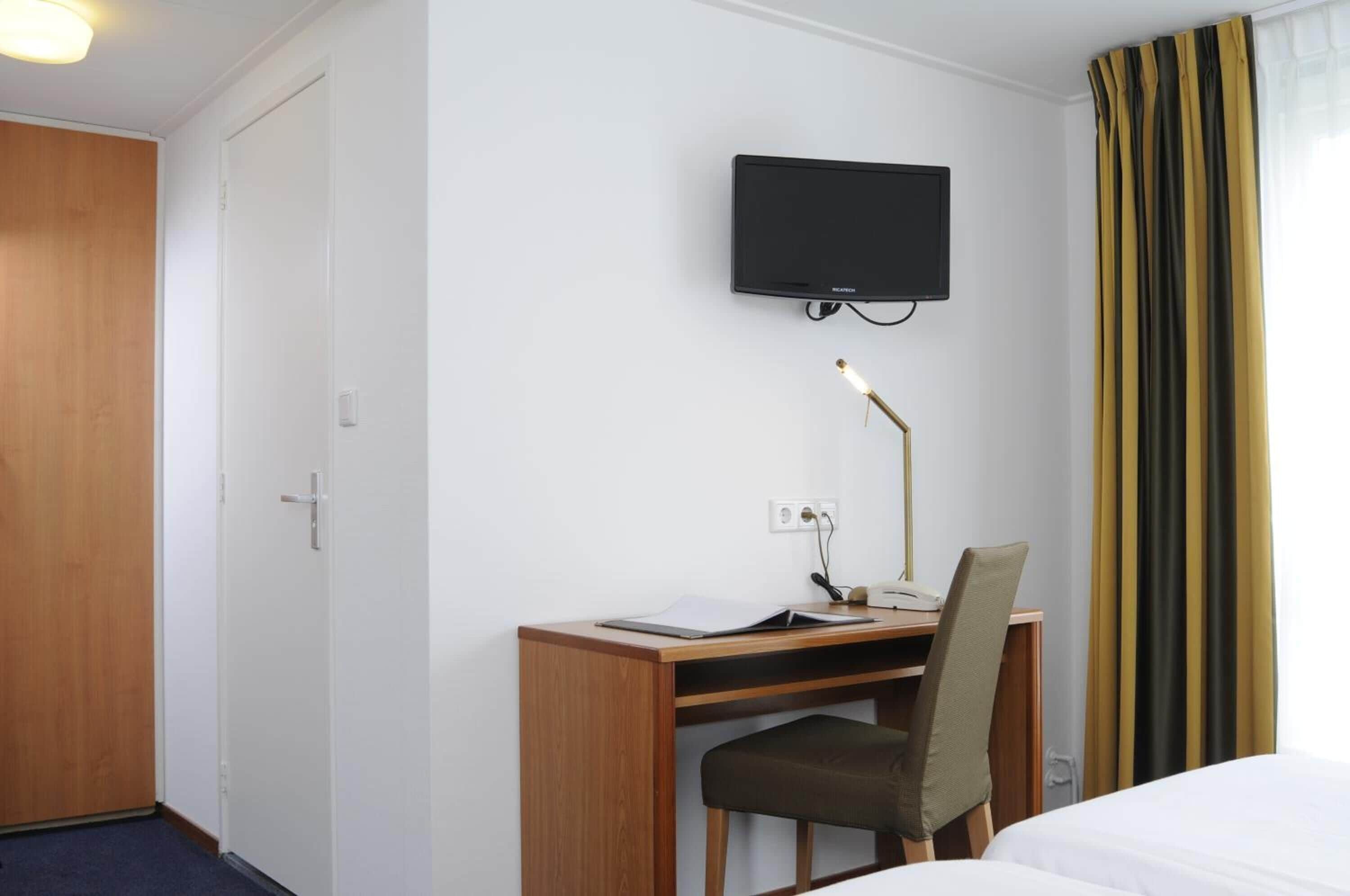 Quarto de hotel com escrivaninha de madeira, uma cadeira, televisão presa na parede e uma cama.