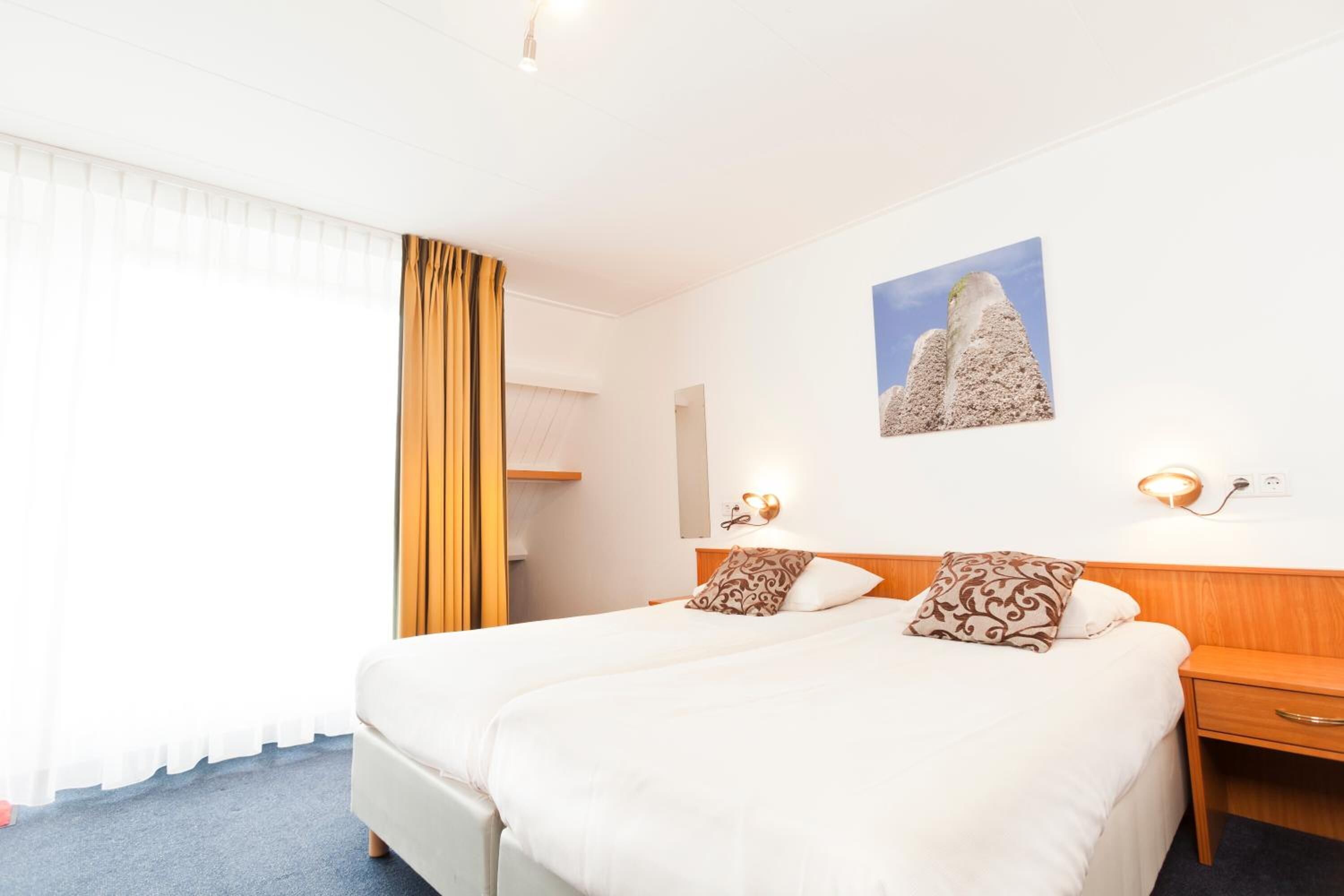 A hotel room with a large bed, two bedside tables, a nightstand, a wall-mounted lamp, and a window with curtains.