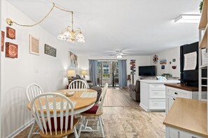 Un coin repas avec une table en bois et des chaises blanches, une cuisine équipée d’un réfrigérateur et d’une cuisinière, et un salon avec un canapé et une télévision.