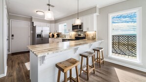 A modern kitchen with a large island, bar stools, pendant lighting, and a view of the ocean through a balcony.
