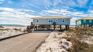 A beachfront house with a deck, a sandy path leading to the entrance, and a view of the ocean.