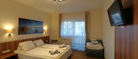 A hotel room with a double bed, a TV mounted on the wall, a painting on the wall, and a window with curtains.