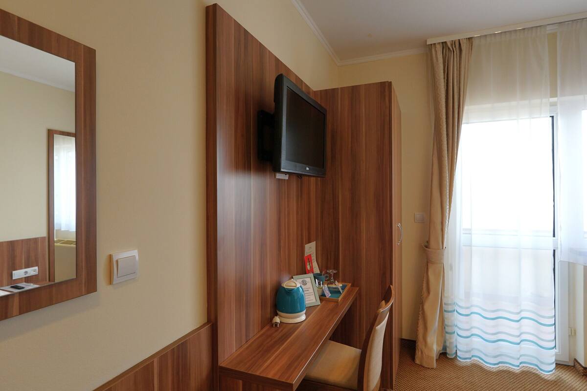 A hotel room with a wooden desk, a television mounted on the wall, a window with curtains, and a mirror on the wall.