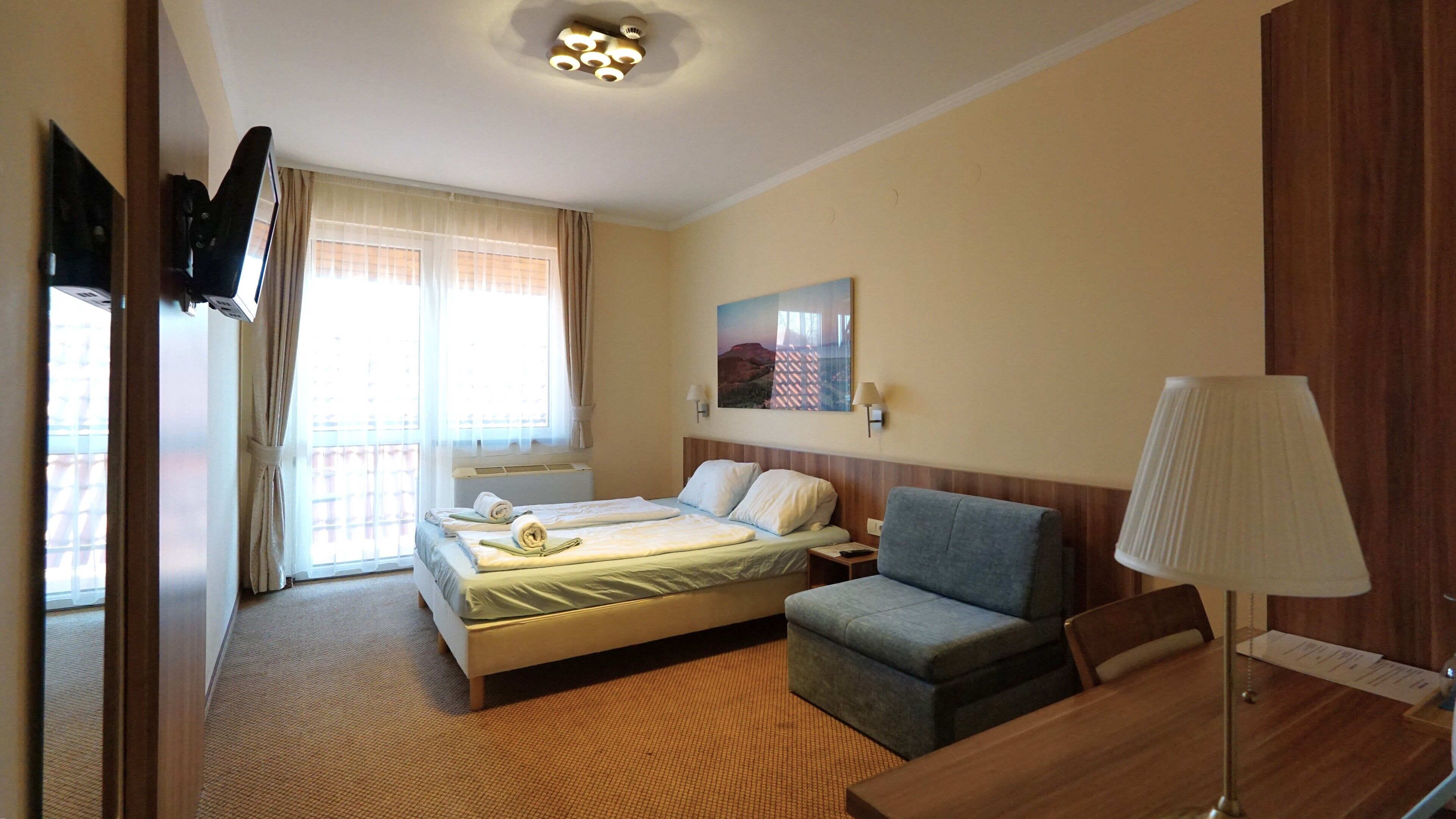 Una habitación de hotel con cama, escritorio, silla, lámpara, un cuadro en la pared y una ventana con cortinas.