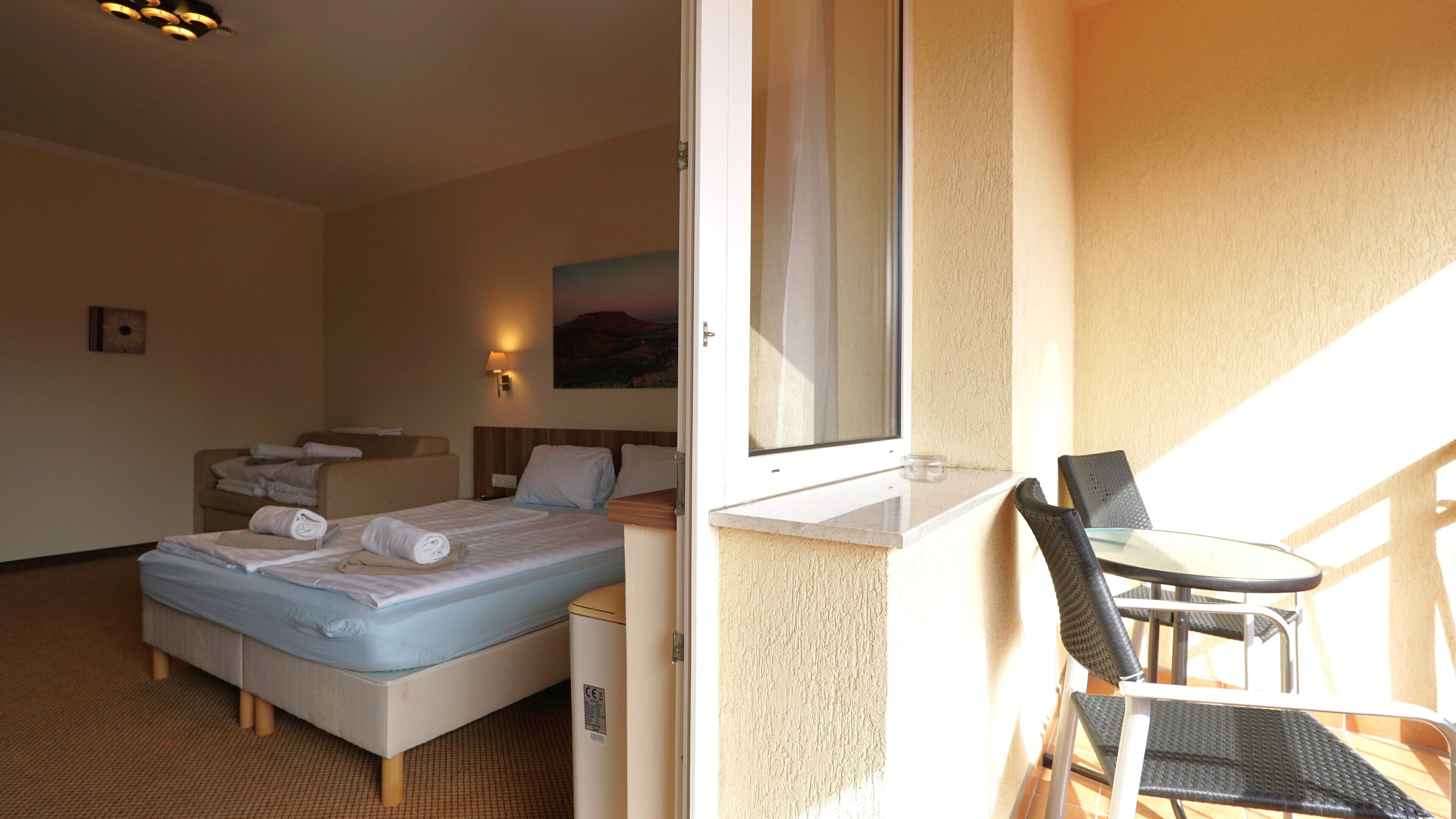 A hotel room with a bed, a small table, a chair, a window, and a painting on the wall.