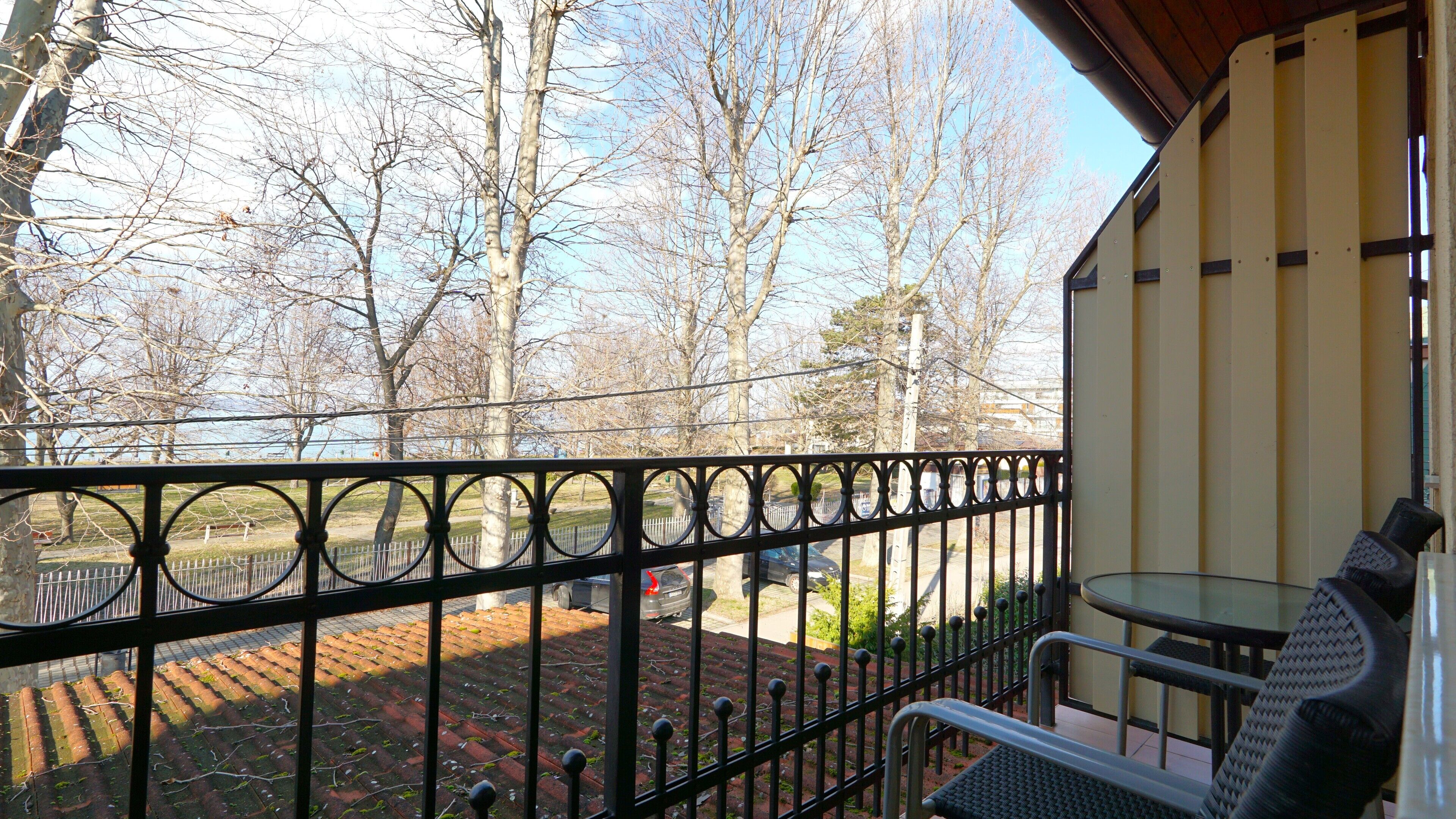 Ein Balkon mit Metallgeländer, einem Tisch und Stühlen. Im Hintergrund sind Bäume und eine gepflasterte Fläche zu sehen.