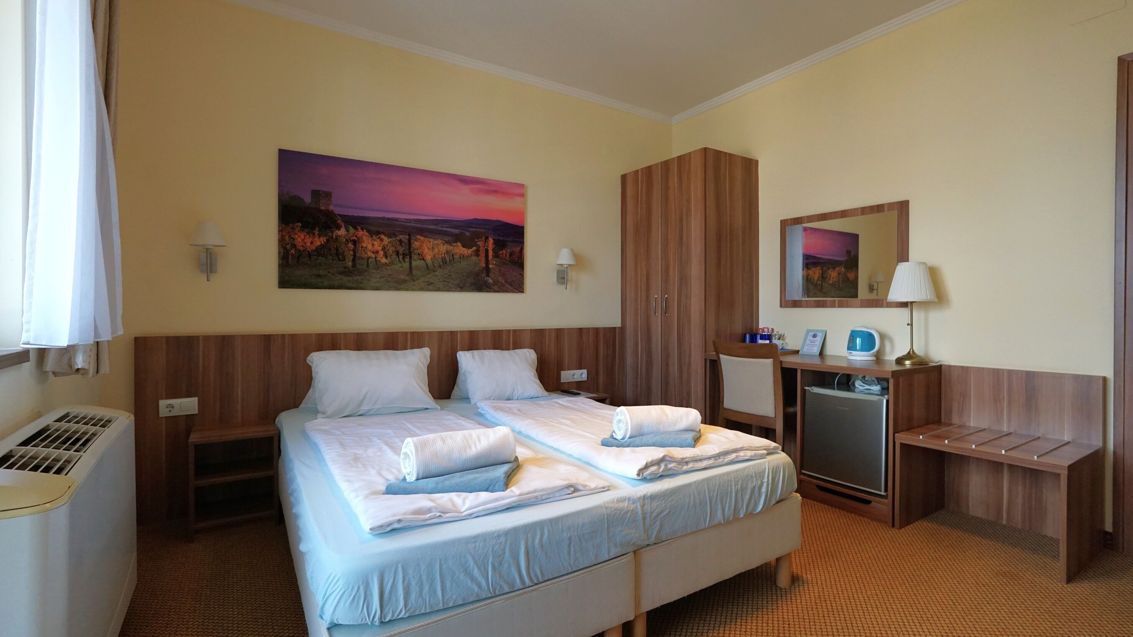 A hotel room with a double bed, a desk with a chair, a refrigerator, and a wall-mounted mirror.
