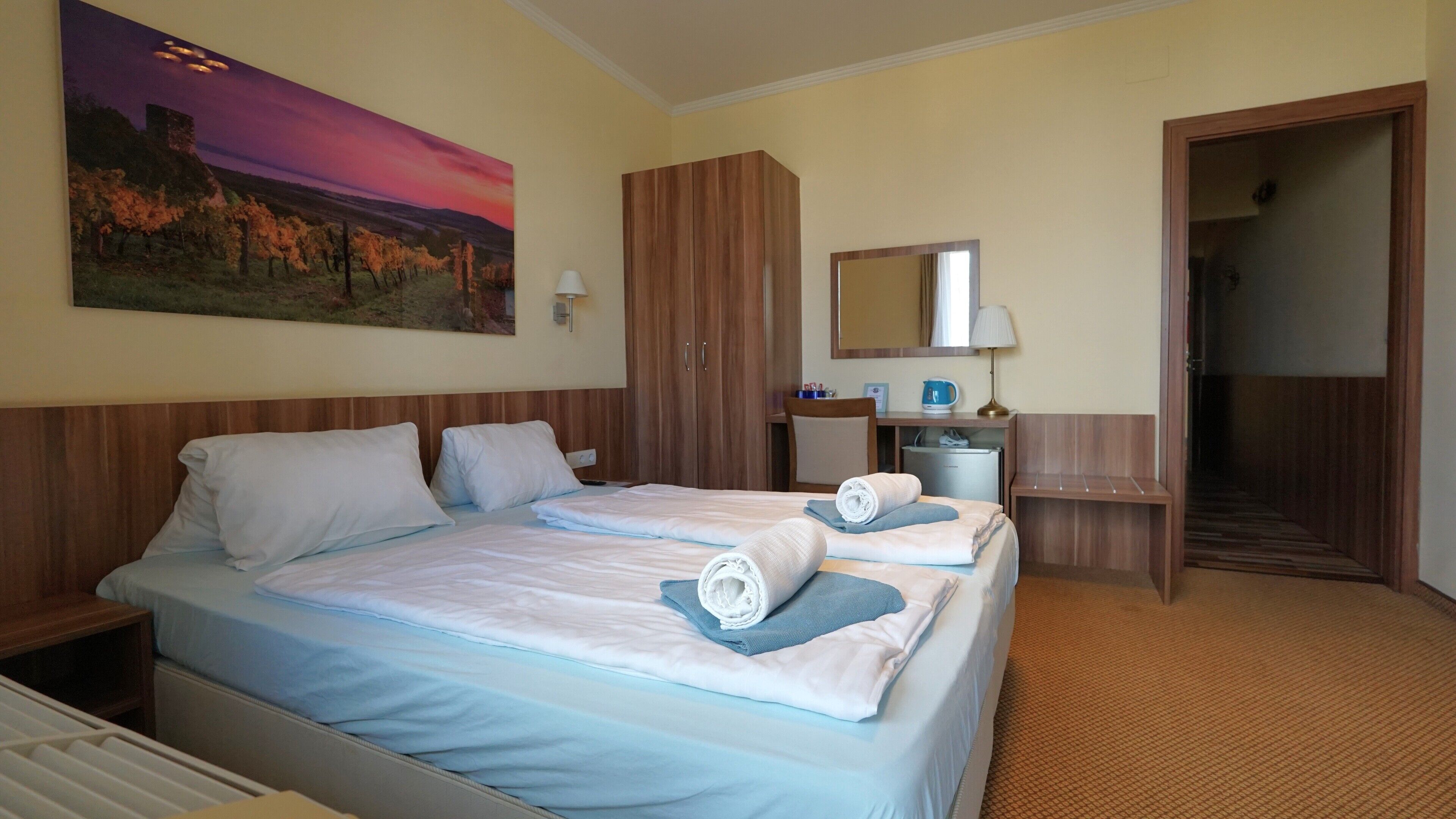 Habitación de hotel con dos camas, un escritorio, una silla, un espejo y un cuadro de un paisaje al atardecer.