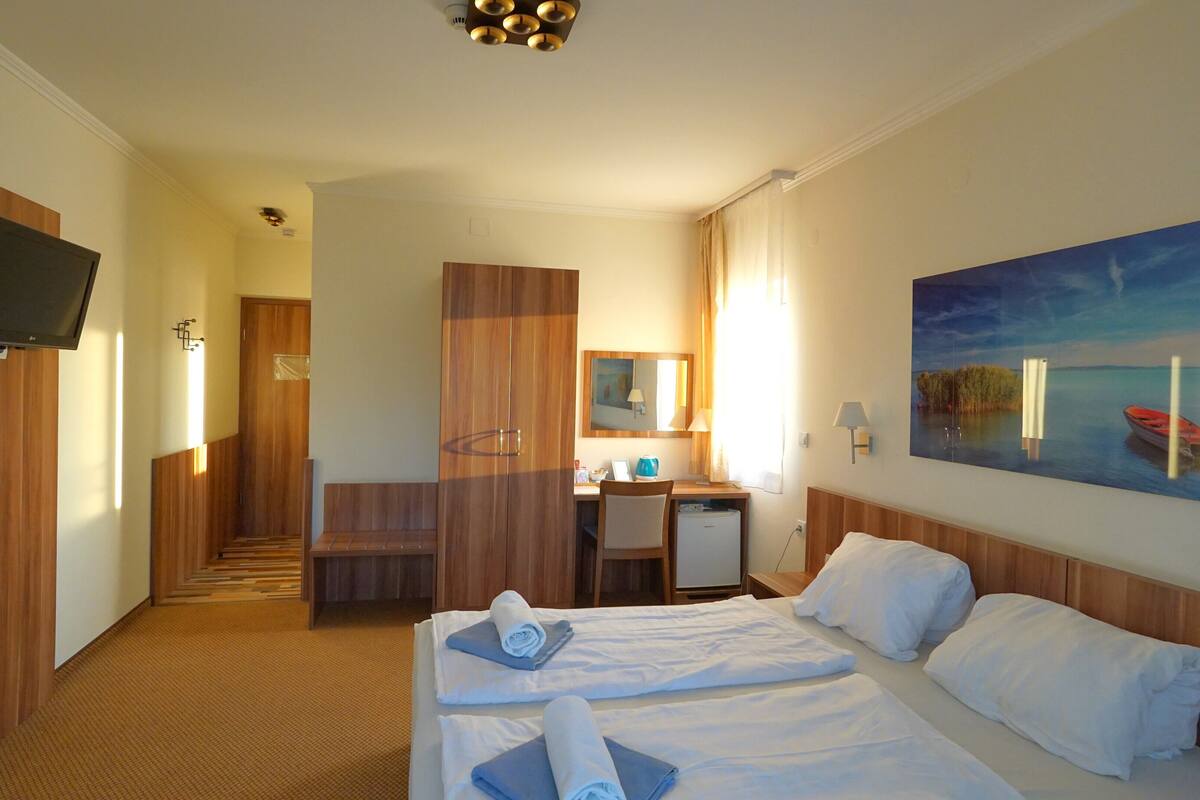 A hotel room with a bed, a desk, a TV, and a painting of a lake.