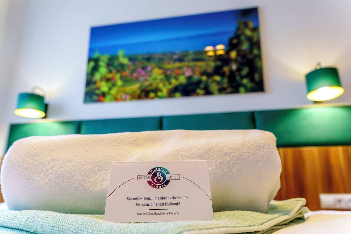 A hotel room with a neatly folded white towel, a sign with the Balaton Colors Beach Hotel logo, and a painting of a landscape on the wall.