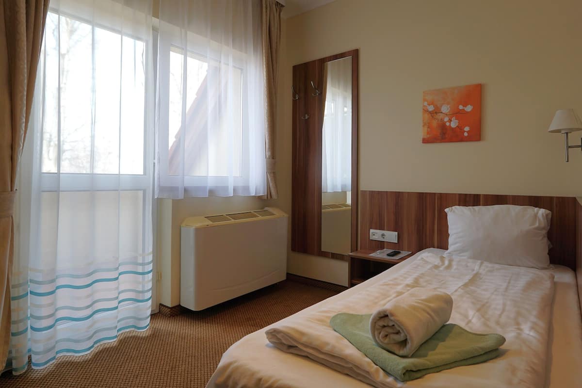 A hotel room with a bed, a bedside table, a mirror, a window with curtains, and an air conditioning unit.
