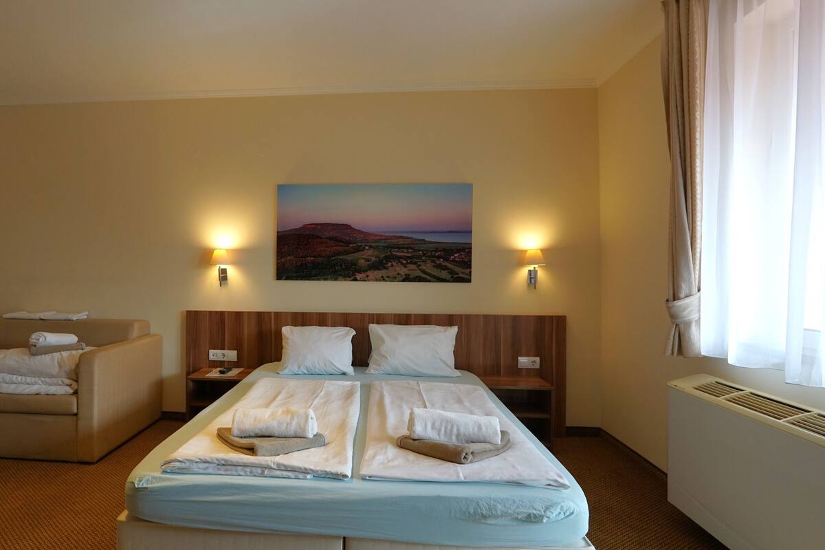 A hotel room with a bed, a sofa, two bedside tables, a wall-mounted lamp, and a painting of a landscape.