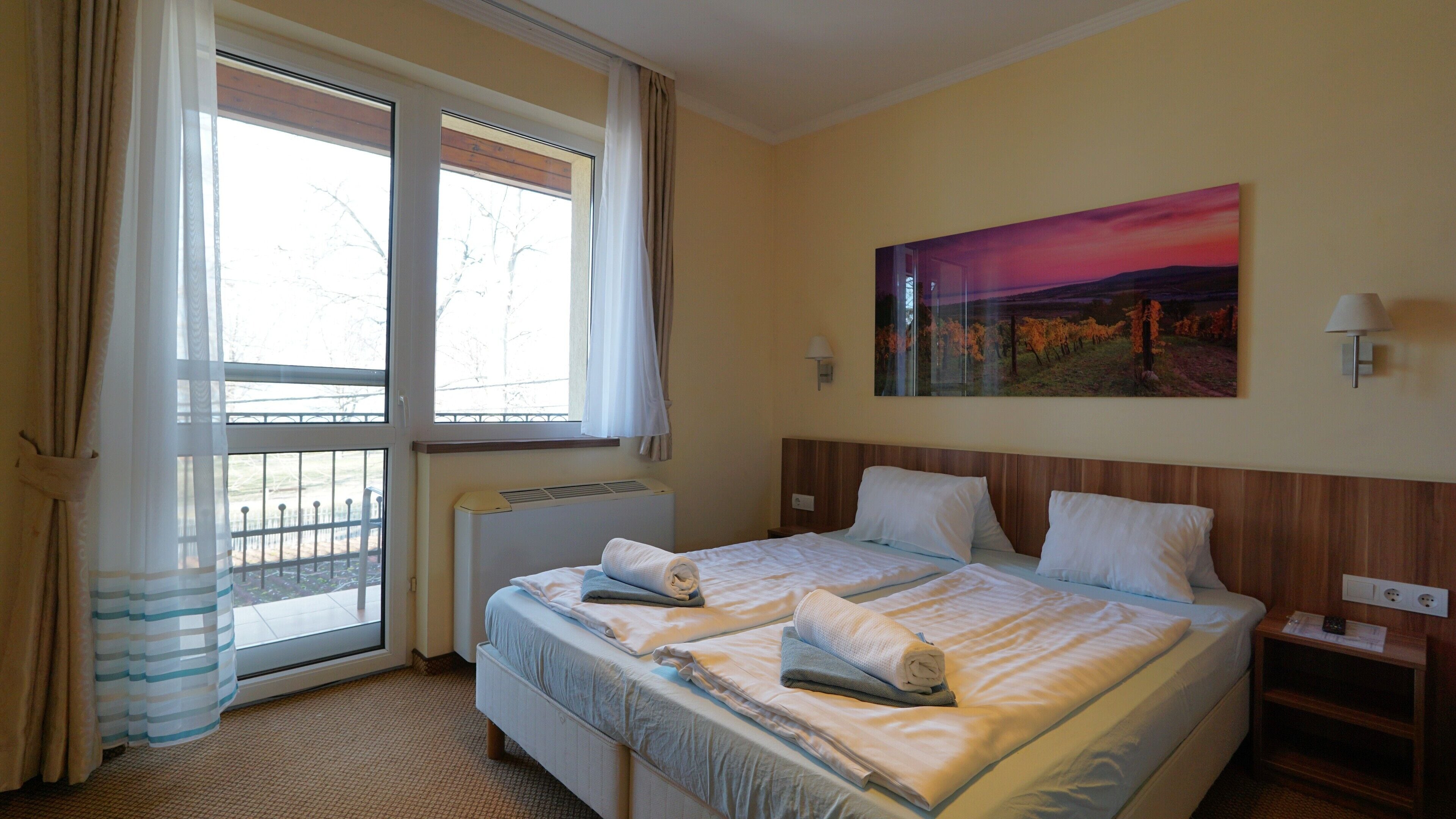 Een hotelkamer met een bed, een raam met gordijnen, een schilderij aan de muur en een nachtkastje met een lamp.