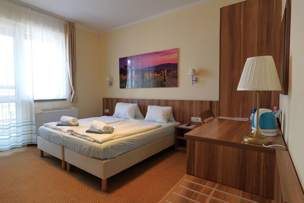 A hotel room with a bed, a desk, a lamp, a painting, and a window with curtains.