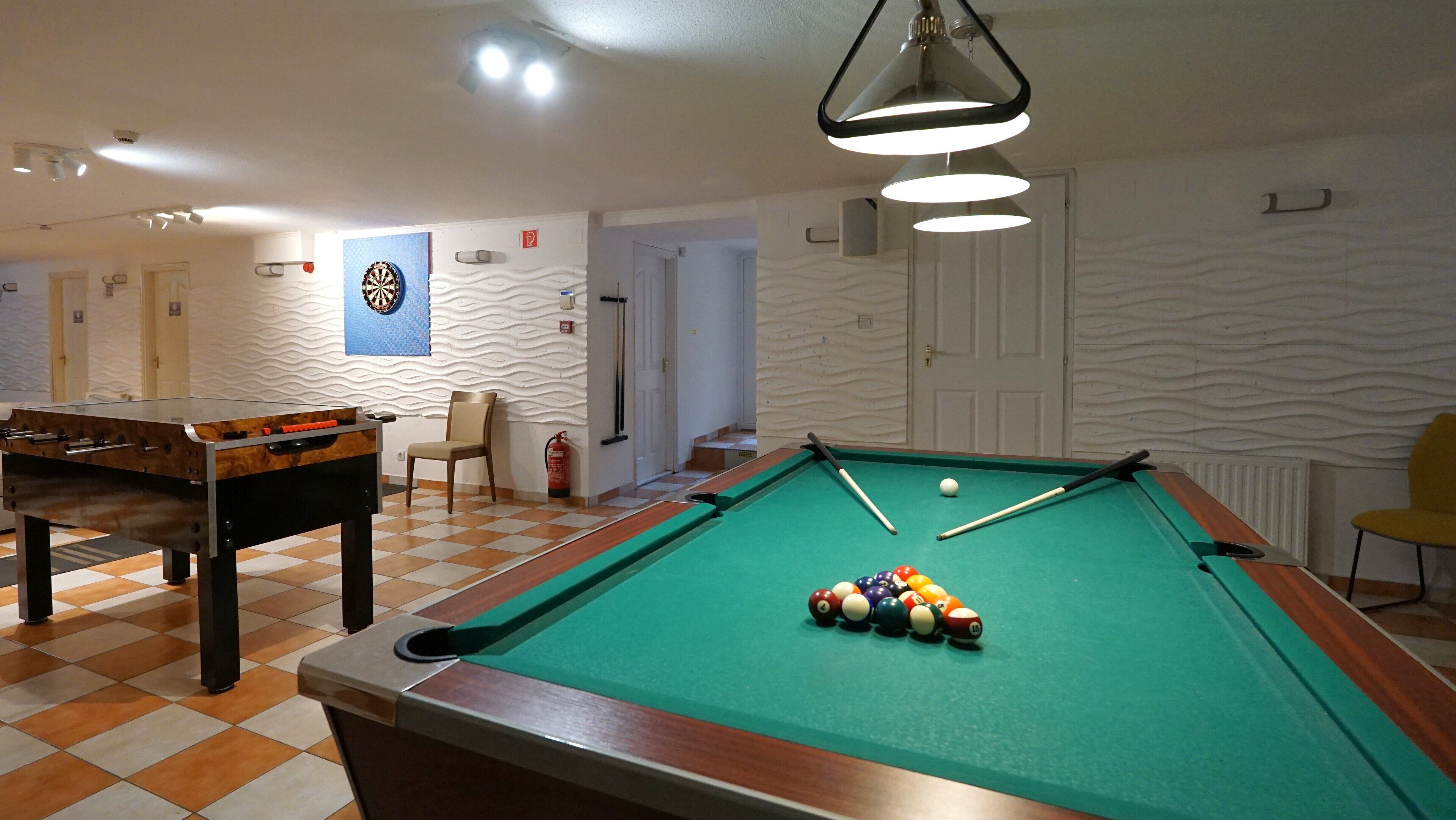 A pool table with billiard balls, a foosball table, and a dartboard in a room with patterned walls and a door in the background.