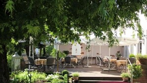 An outdoor patio area with tables and chairs, surrounded by greenery and a building with a sign. - Landgasthof Hirsch (Remshalden)