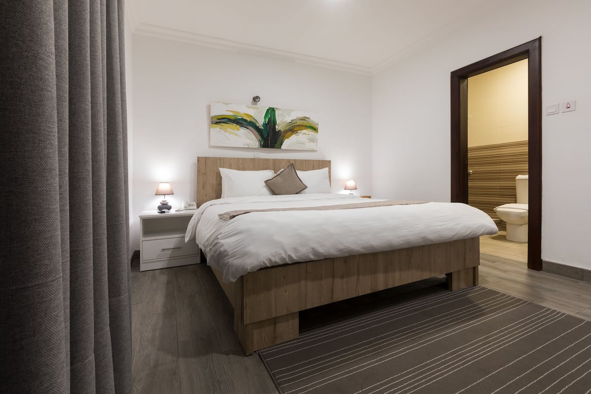 Ein modernes Schlafzimmer mit einem hölzernen Bett, weißer Bettwäsche, einem Nachttisch mit Lampe und einem Gemälde an der Wand.
