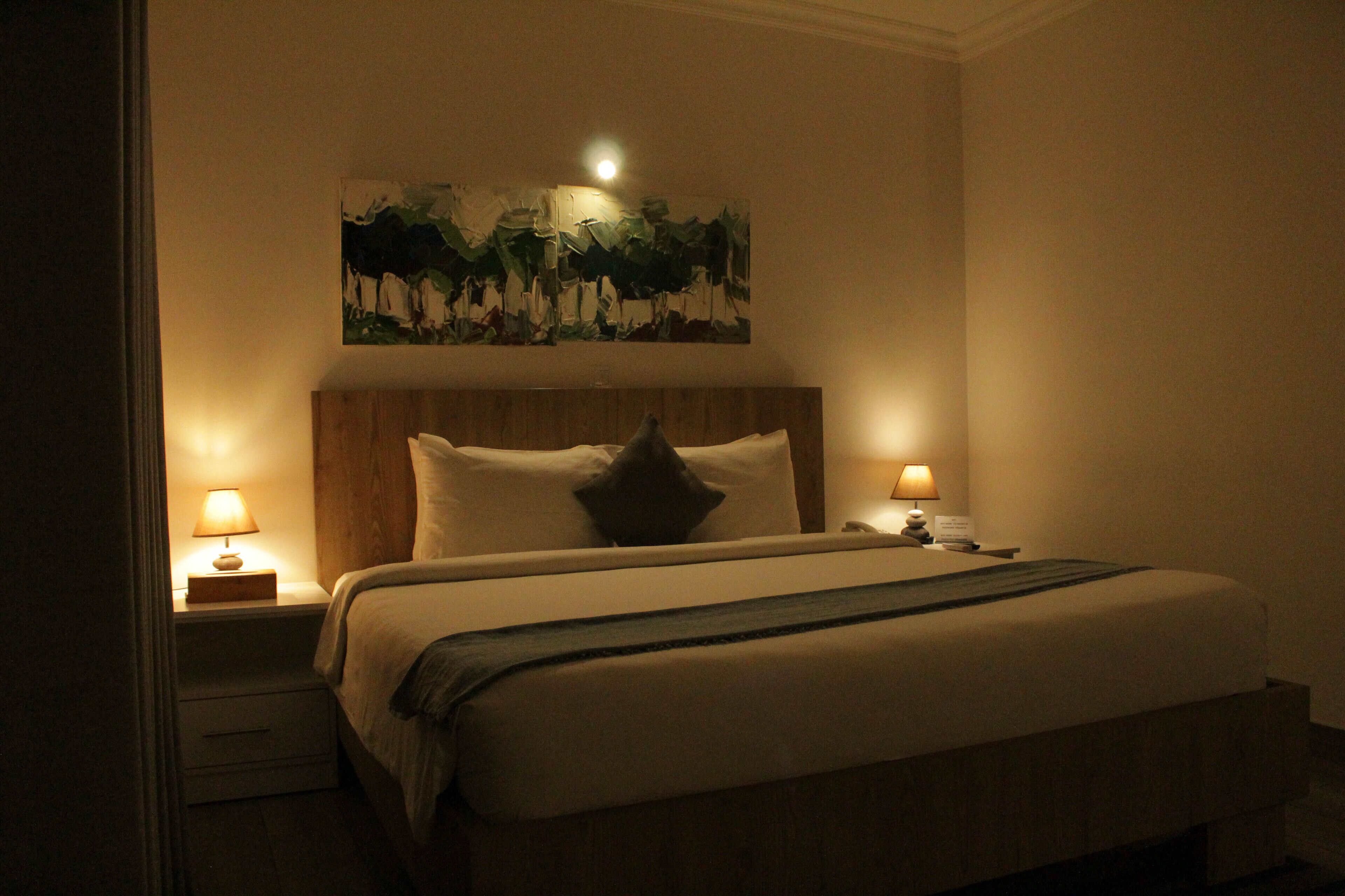Habitación de hotel con una cama grande, dos lámparas de noche, una mesita de noche y un cuadro colgado en la pared.