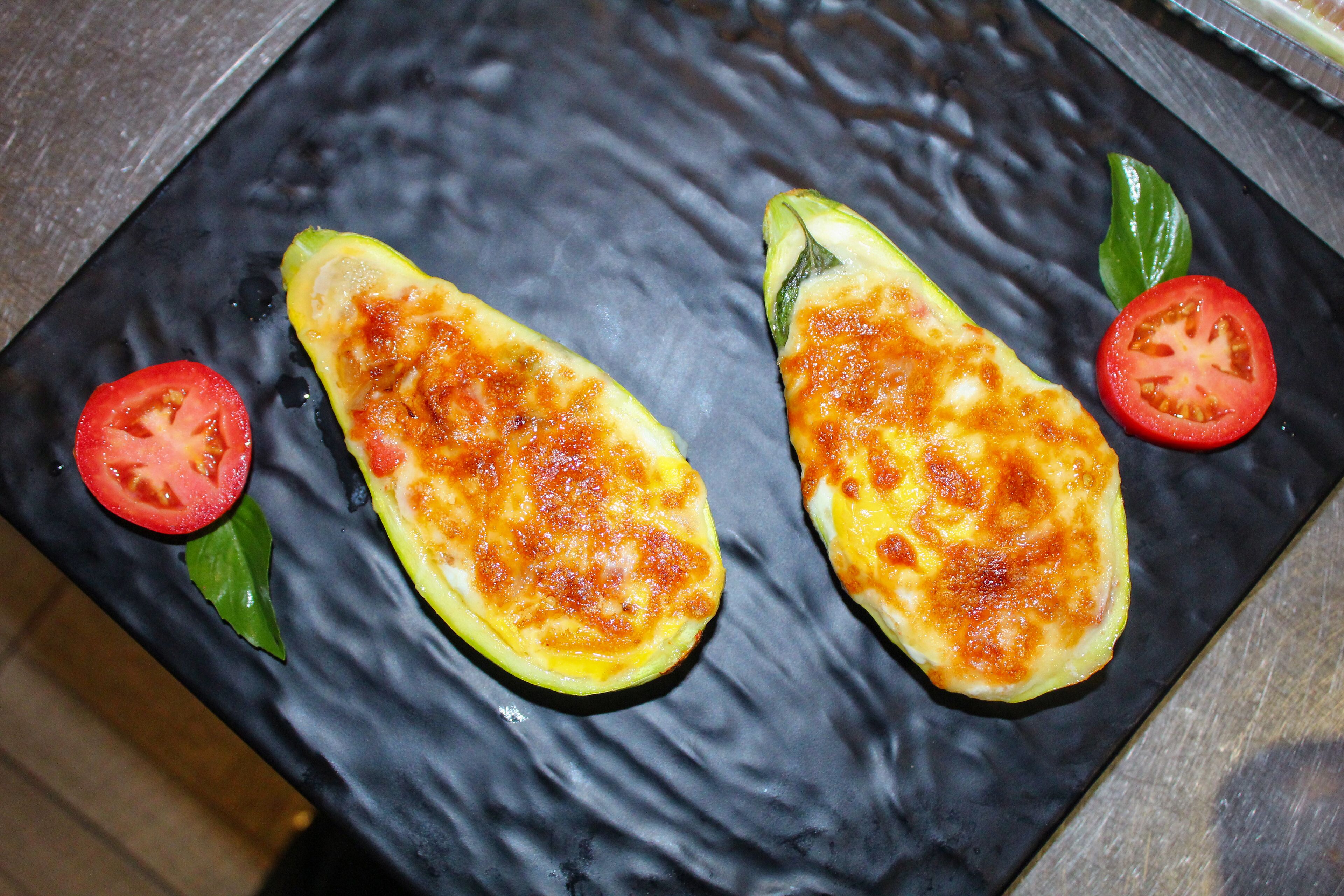 Two grilled zucchini halves on a black plate with tomato slices and basil leaves.