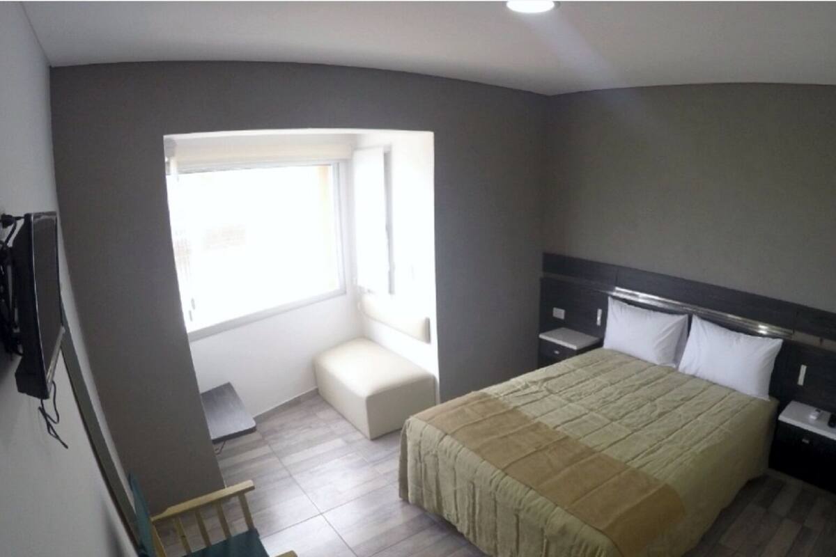 A bedroom with a bed, a bedside table, a nightstand, a window, and a small seating area.
