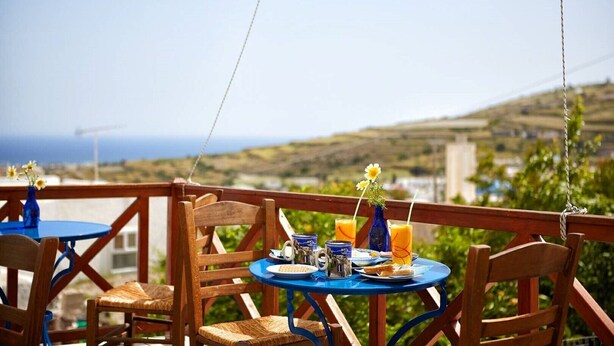 A terrace with wooden chairs and tables, a vase with flowers, and a view of the sea and hills. - Santorini-treasures Rockside Villa traditional (Emporio)