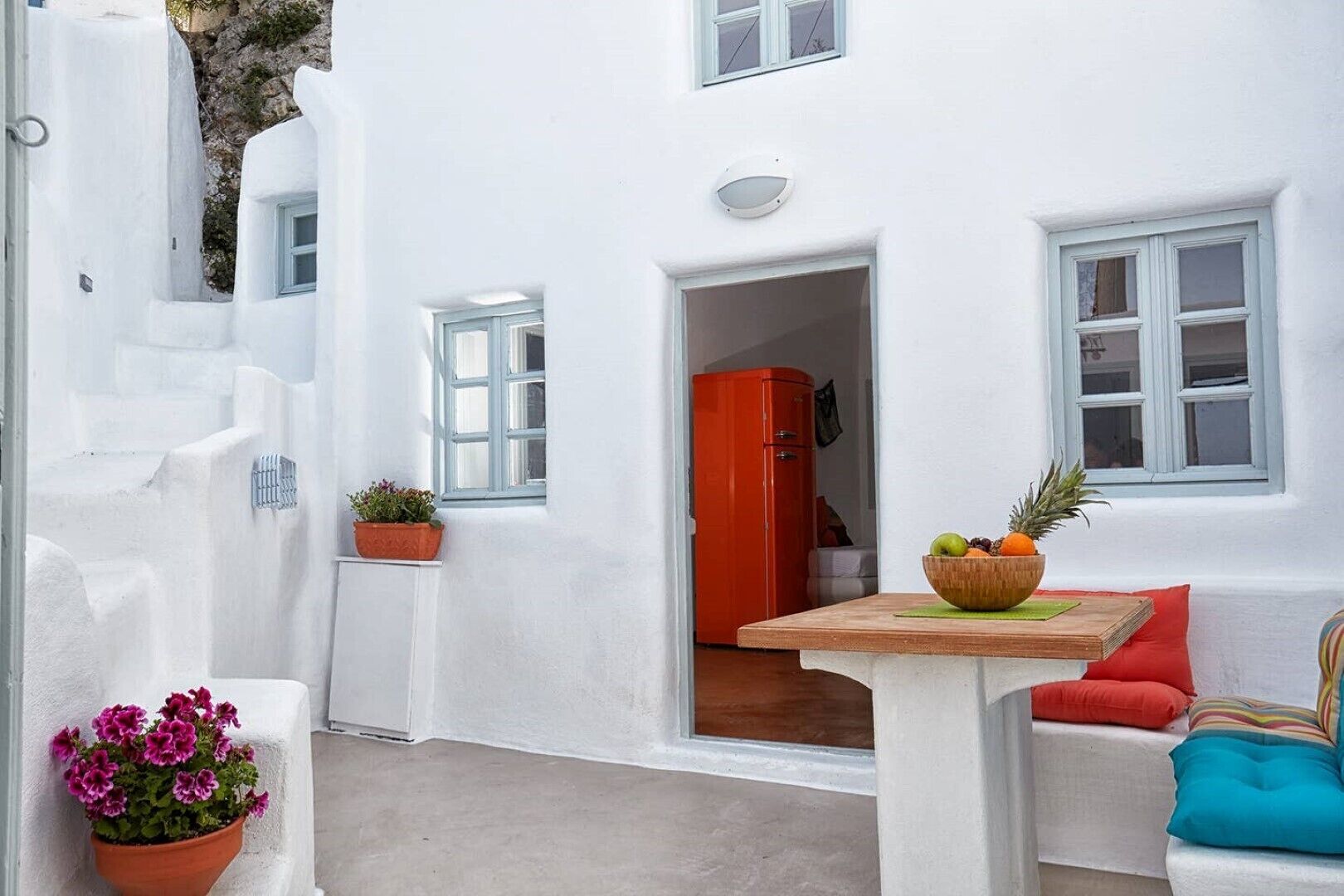 A white-walled building with a small table and chairs, a potted plant, and a red door.