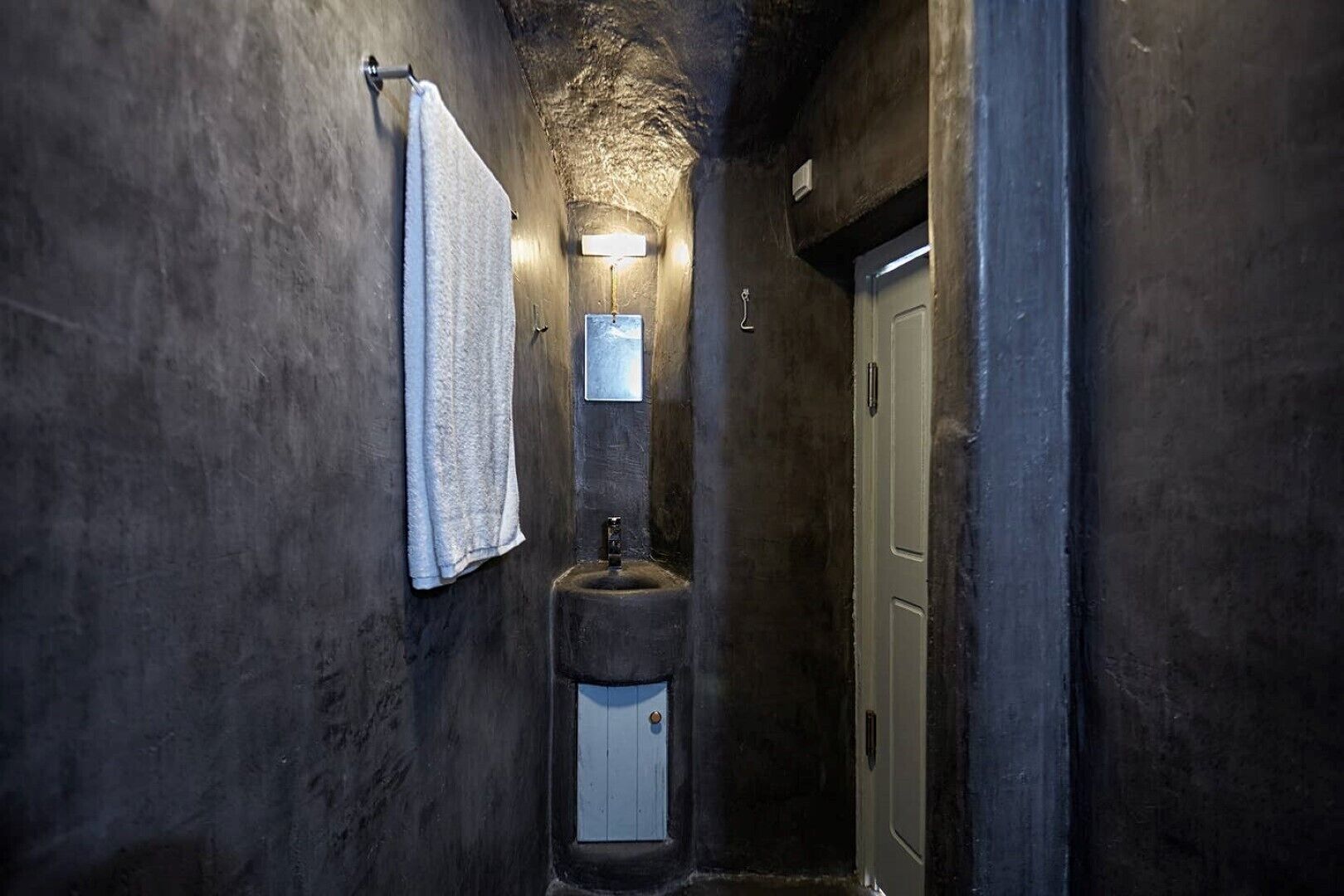 Ein schmales, schlicht beleuchtetes Badezimmer mit rauer Wandstruktur, einem Einzelwaschbecken und einem Handtuch an einer Metallstange.