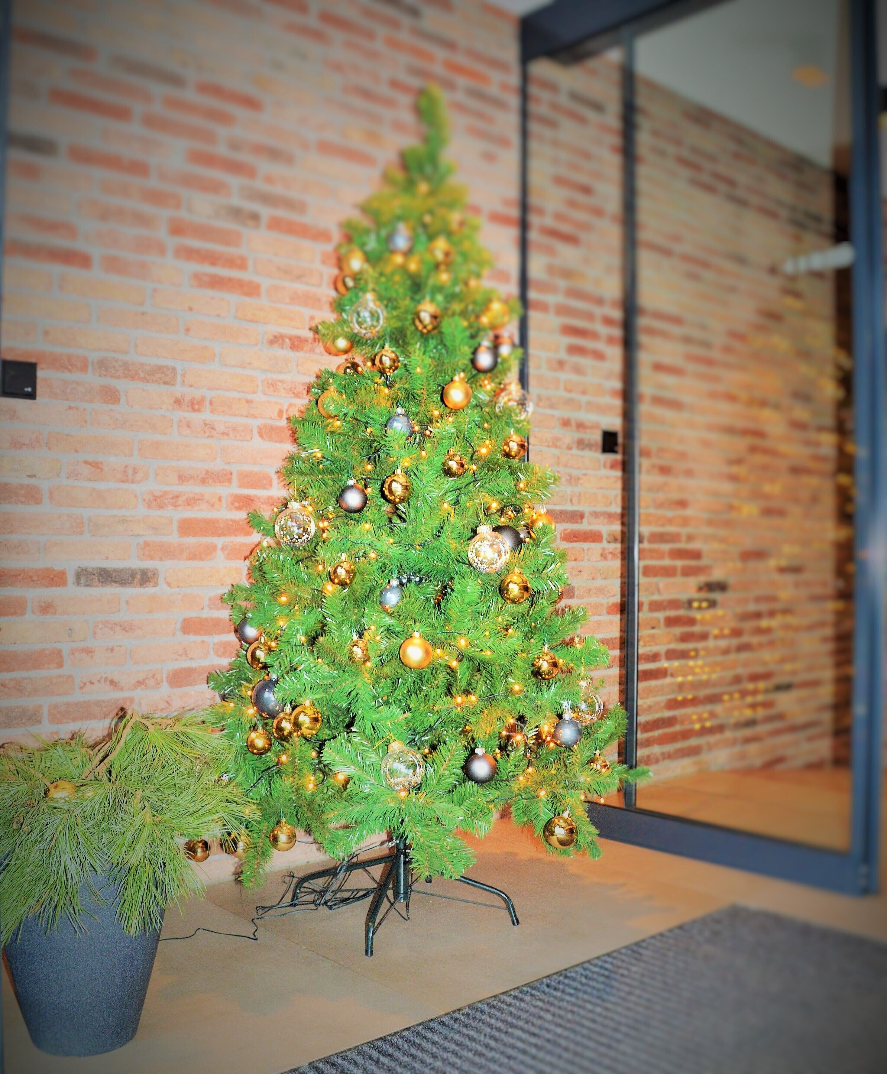 A Christmas tree with gold and silver ornaments in front of a brick wall.