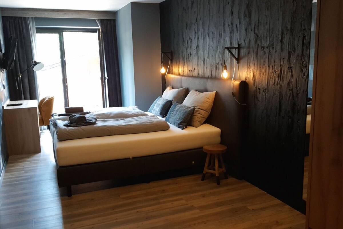 Ein modernes Schlafzimmer mit Holzboden, einem Bett mit vielen Kissen, einem kleinen Hocker und einer Wandleuchte.