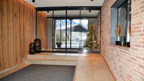 A modern lobby with a wooden wall, a glass door leading outside, a Christmas tree, and a brick wall with a window. - Hotel Herz3 (Hollersbach im Pinzgau)