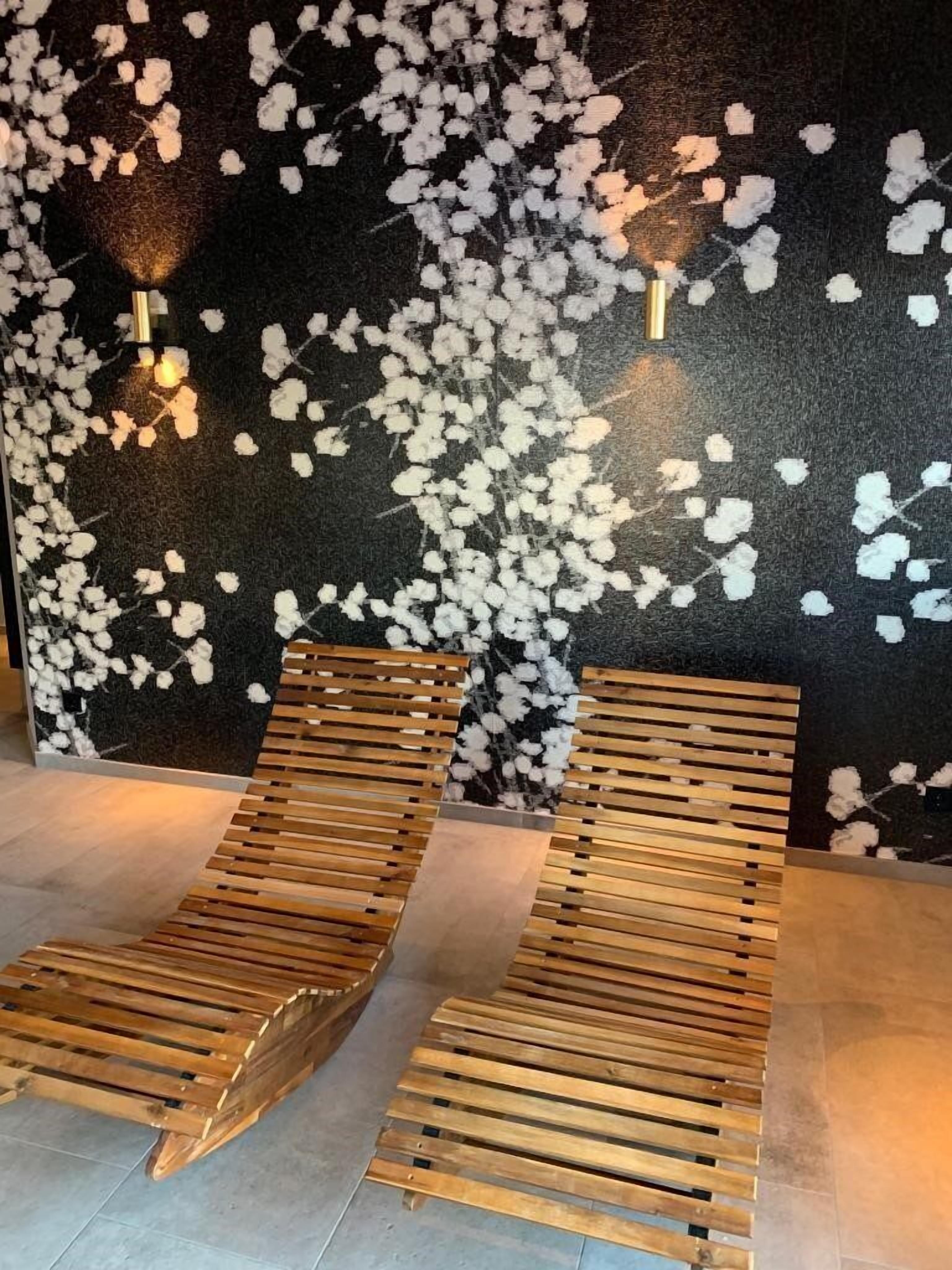 Een kamer met twee houten lounge fauteuils, een donkere wand met textuur en witte bloemmotieven, en twee wandlampen.
