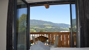 A room with a bed, a view of a balcony with chairs, and a wooden deck. - Hotel Herz3 (Hollersbach im Pinzgau)
