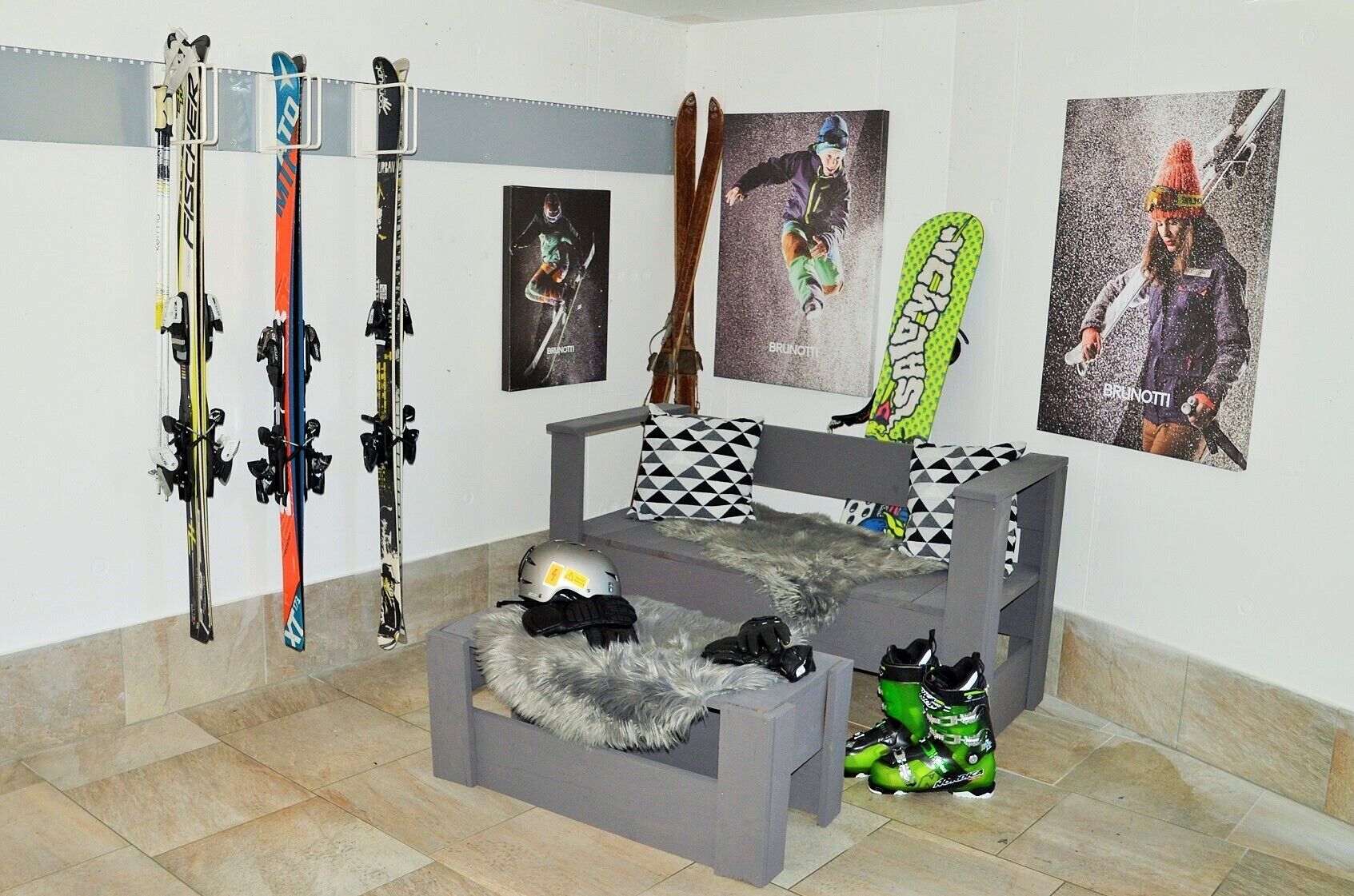Una habitación con un sofá gris, un plaid de piel y un casco sobre una mesa. Hay esquíes y una tabla de snowboard apoyados contra la pared.