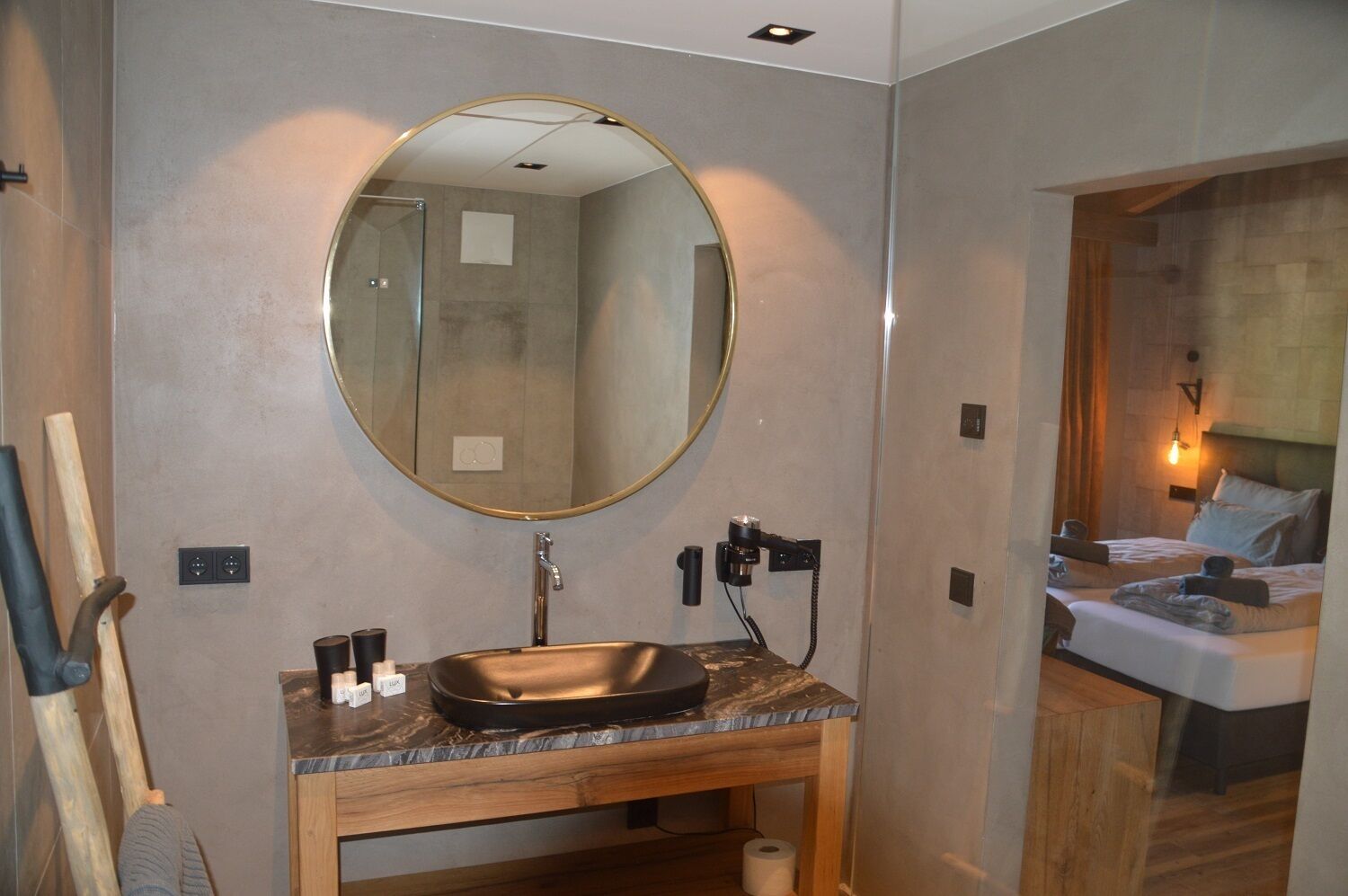 A modern bathroom with a round mirror, a sink, and a shower. There is a wooden bench and a towel rack.