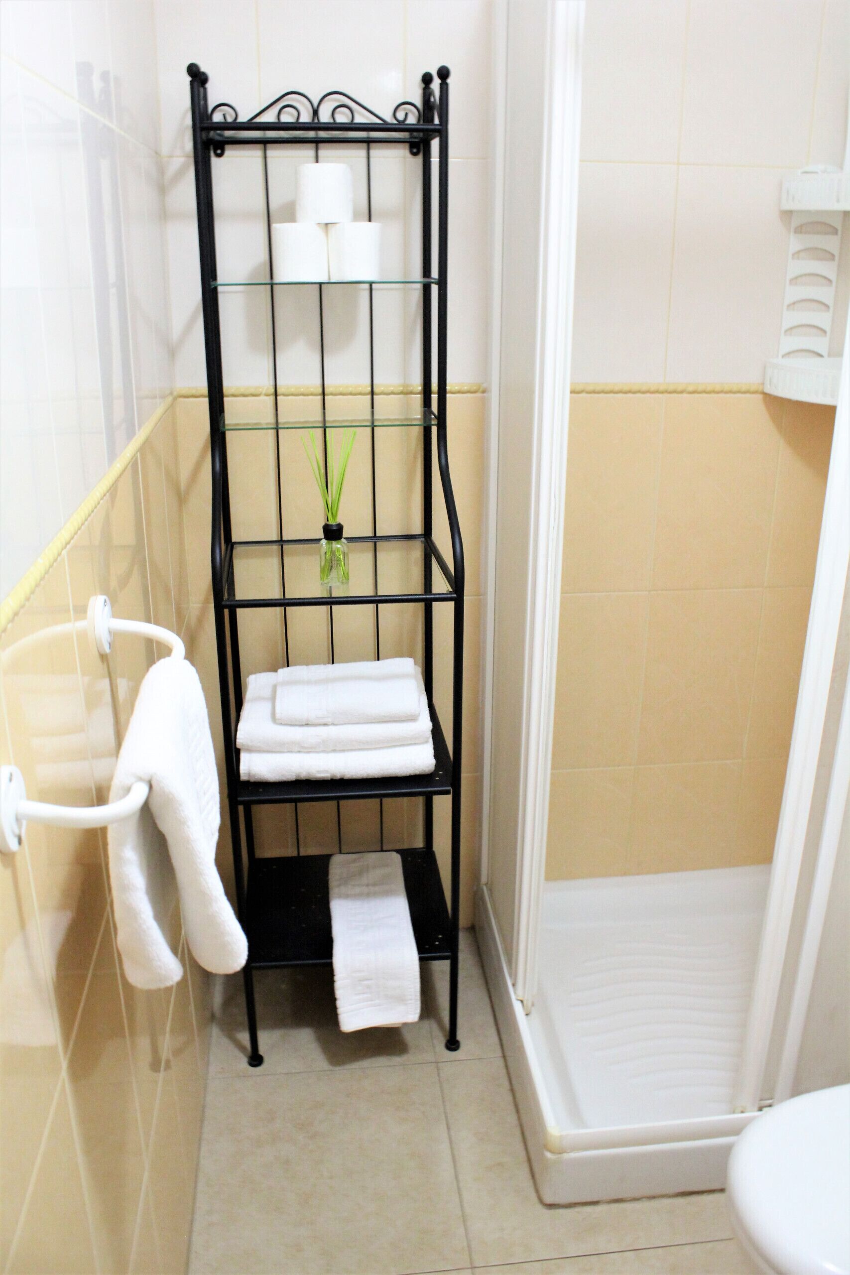 A bathroom with a black metal rack holding towels and a small plant, a white toilet, and a shower area.