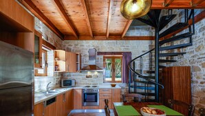 Una cocina rústica con gabinetes de madera, un refrigerador de acero inoxidable, una estufa y un fregadero. Se aprecia una mesa de comedor con un mantel individual verde y un tazón de frutas. Se ve una escalera de caracol al fondo.