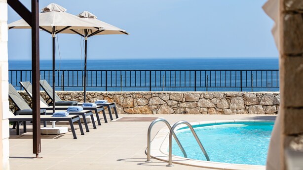 Un área junto a la piscina con sillones de descanso y sombrillas, un muro de piedra y vistas al océano.