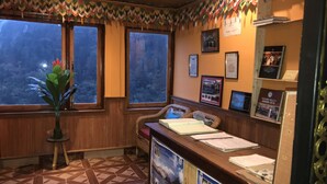 A reception area with a wooden desk, two wicker chairs, a potted plant, and a patterned floor. - Rufina Blue Pine (Chungthang)