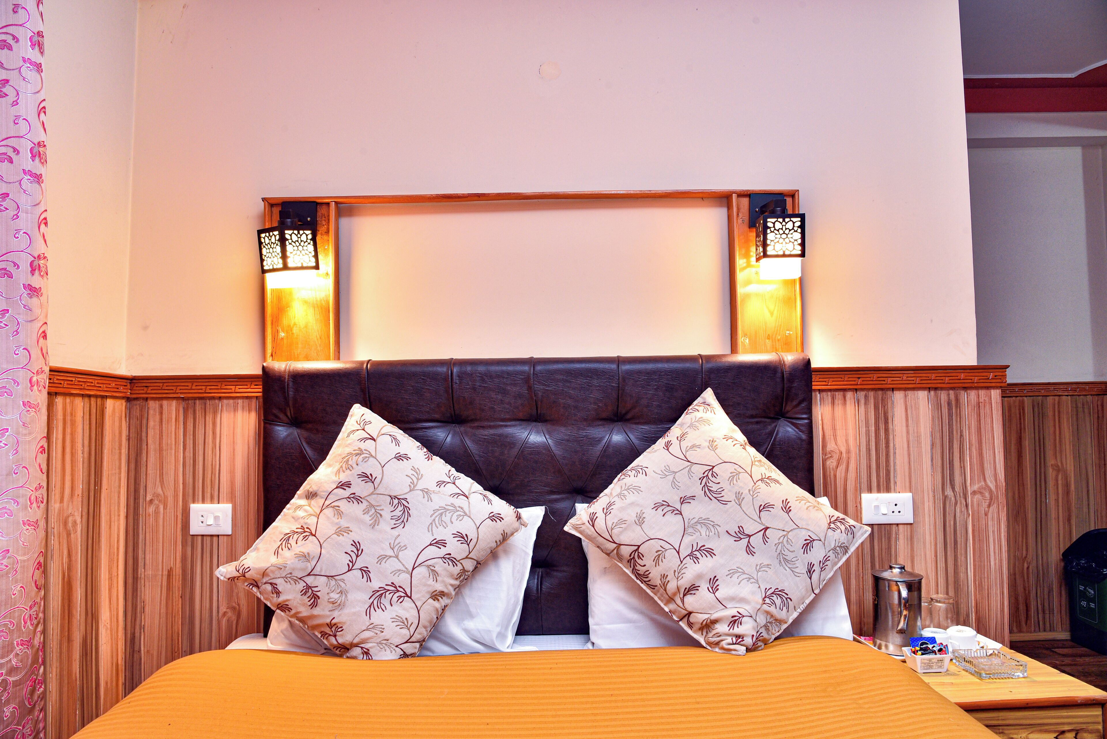 Habitación de hotel con un cabecero de cuero marrón, dos apliques decorativos, una mesita de noche de madera con una cafetera y una cama con almohadas estampadas.