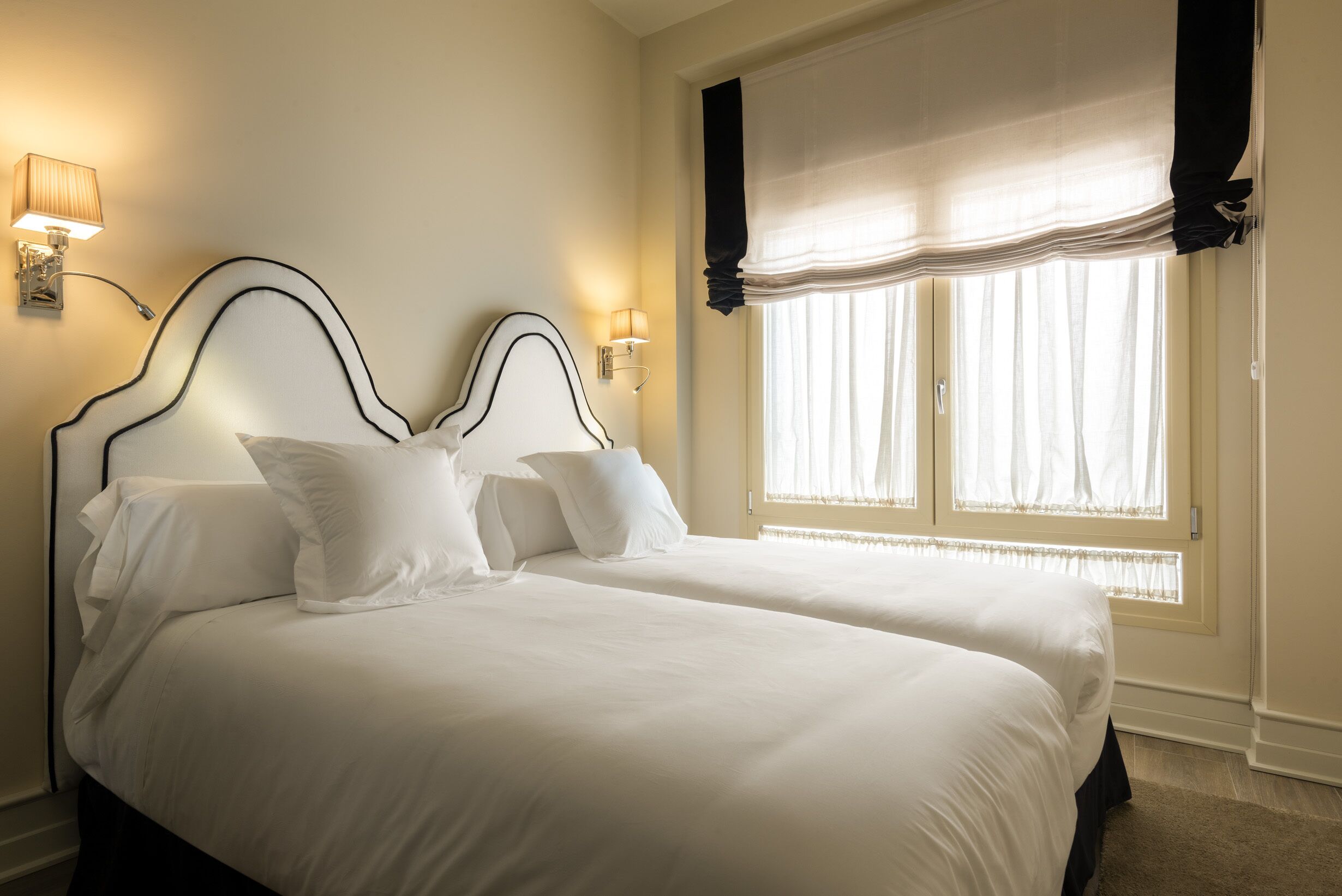 Ein Hotelzimmer mit zwei Betten, einem Fenster mit leichtem Vorhang und Wandleuchten.