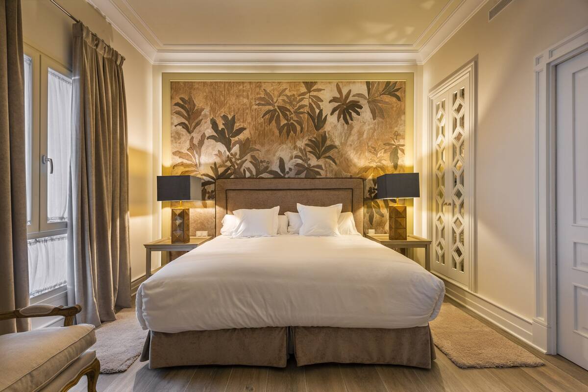 Een groot bed met witte lakens, twee nachtkastjes met lampen, een groot bloementapijt aan de muur en een raam met gordijnen.