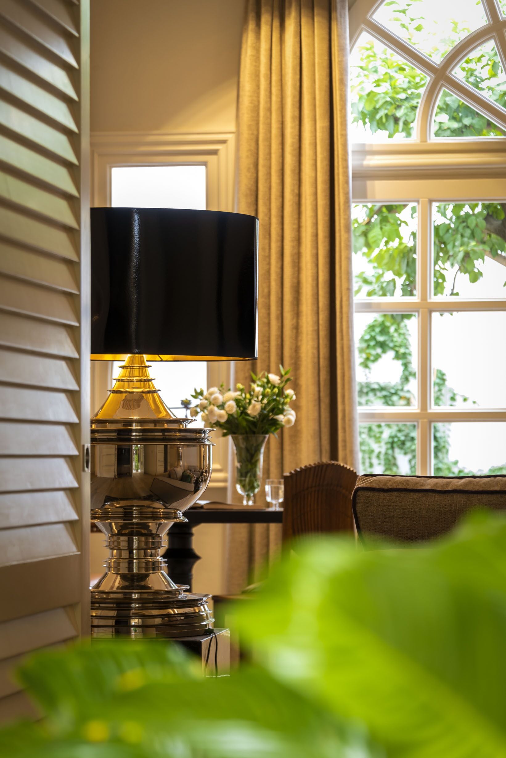 A lamp with a black shade, a vase with flowers, and a window with greenery outside.