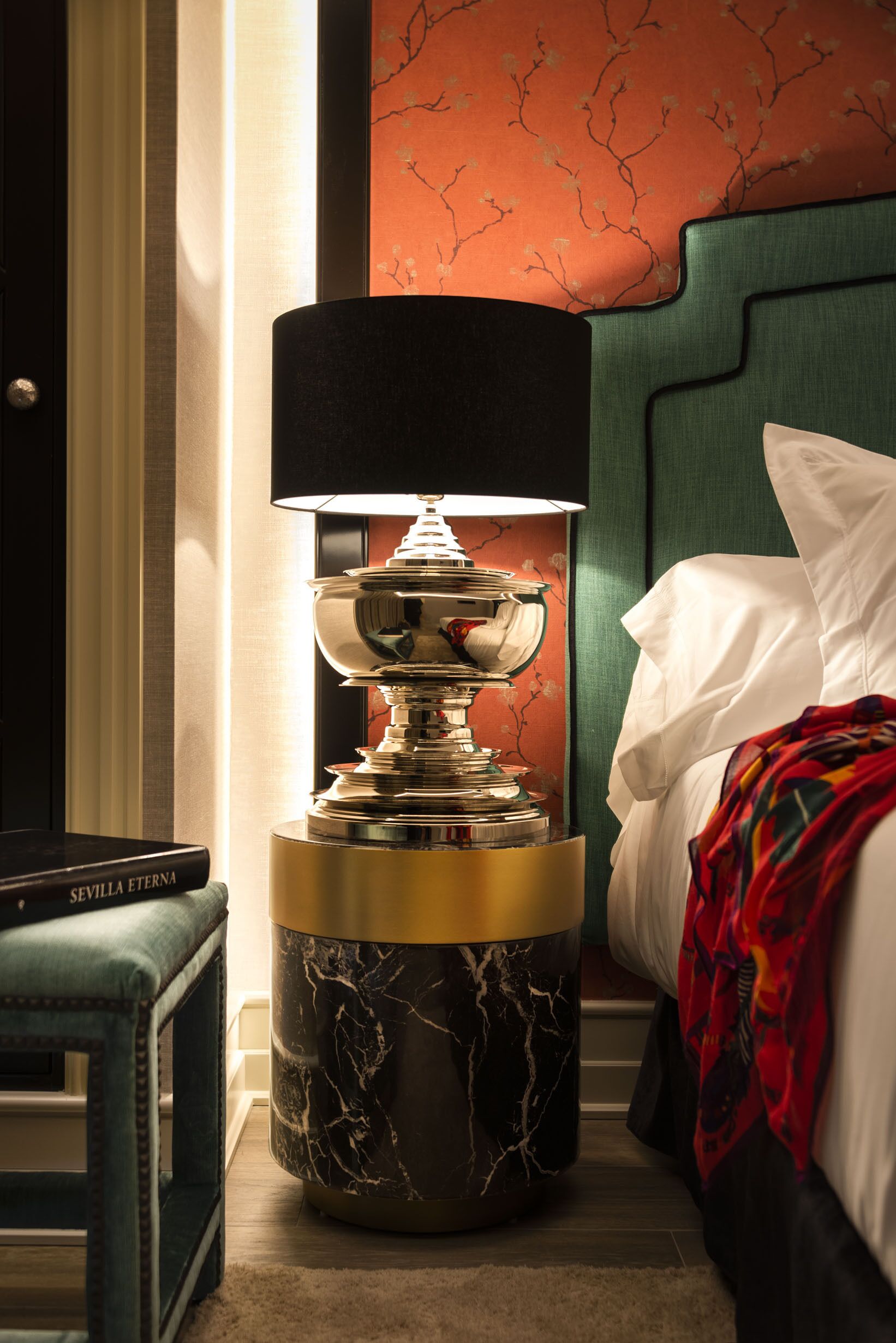 A modern hotel room with a marble-patterned floor lamp, a green upholstered headboard, and a neatly made bed with a white pillow and a colorful throw blanket.