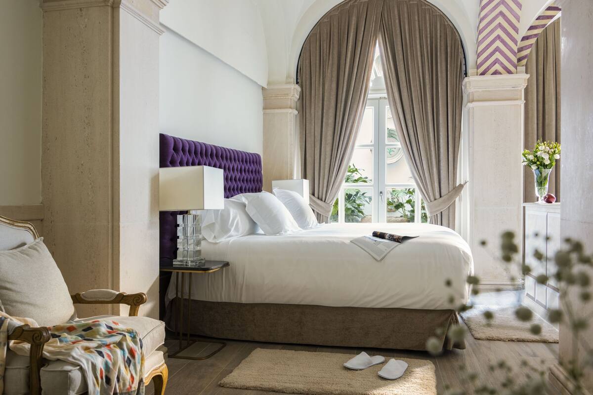 Een slaapkamer met een groot bed, een nachtkastje, een stoel en een raam met gordijnen.
