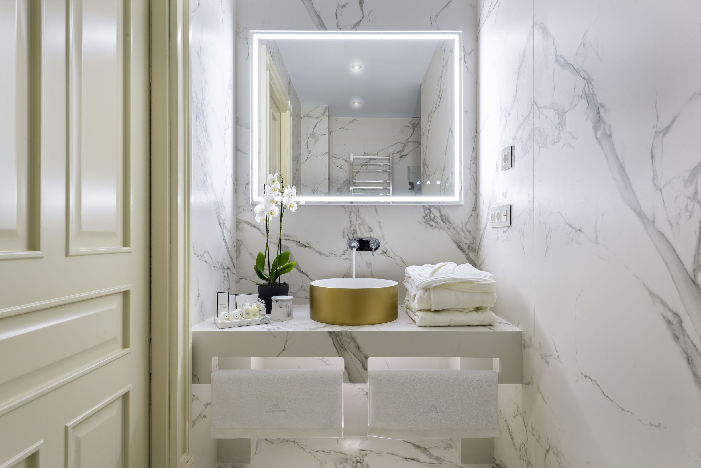 A bathroom with a marble wall, a gold sink, and a mirror with LED lighting.