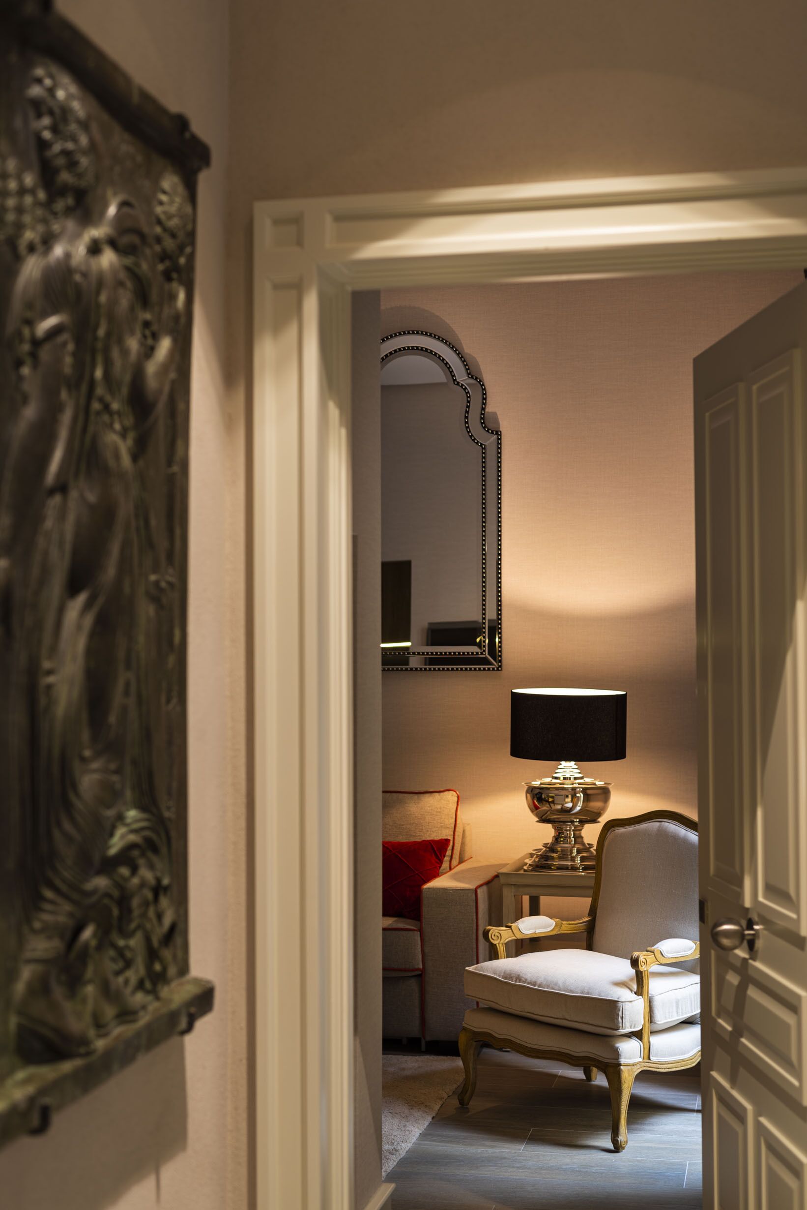 A hallway with a door, a chair, a table with a lamp, and a mirror on the wall.